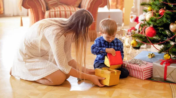 Симпатичный 3-летний мальчик сидит под елкой и смотрит внутрь подарочных коробок — стоковое фото