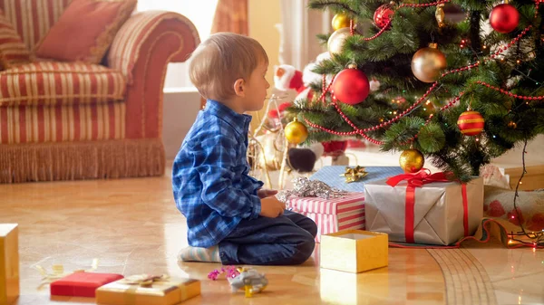 Niño de 3 años sentado bajo el árbol de Navidad y mirando regalos y regalos — Foto de Stock