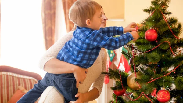 Mulher bonita segurando seu filho pequeno e ajudando-o a decorar a árvore de Natal — Fotografia de Stock