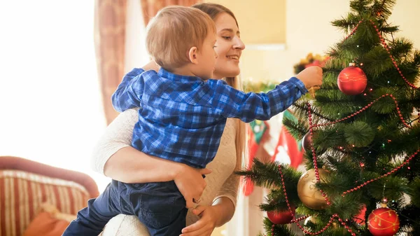 Retrato de una hermosa madre joven abrazando a su niño pequeño y decorando el árbol de Navidad —  Fotos de Stock