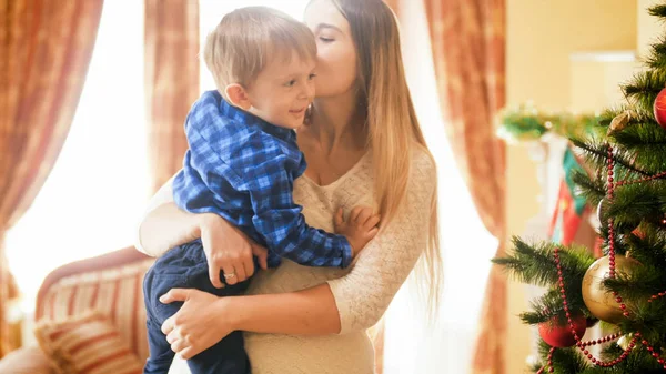 Retrato de madre joven besando a su hijo pequeño junto al árbol de Navidad en la sala de estar —  Fotos de Stock