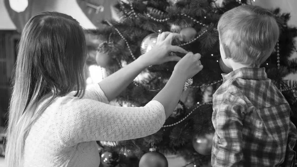 Schwarz-Weiß-Porträt eines kleinen Jungen, der Mutter beim Schmücken des Weihnachtsbaums im Wohnzimmer zusieht — Stockfoto