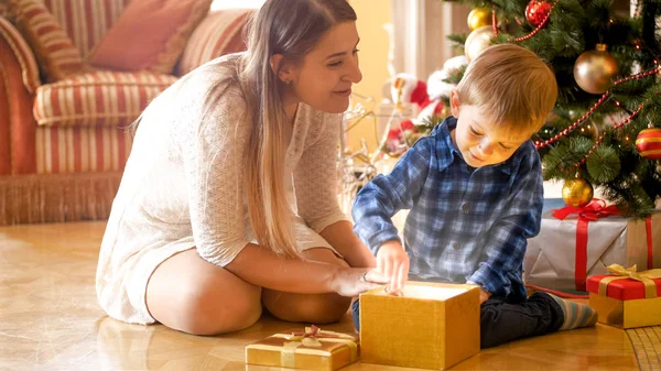Маленький мальчик сидит с мамой под елкой и открывает волшебную светящуюся подарочную коробку — стоковое фото