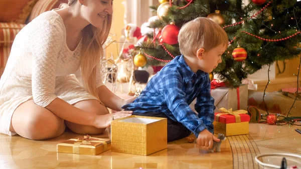 Pequeño niño sentado en el suelo con una hermosa madre joven bajo el árbol de Navidad — Foto de Stock