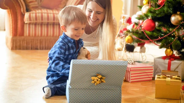Porträtt av leende glad pojke sitter under julgranen och öppna presentaskar — Stockfoto