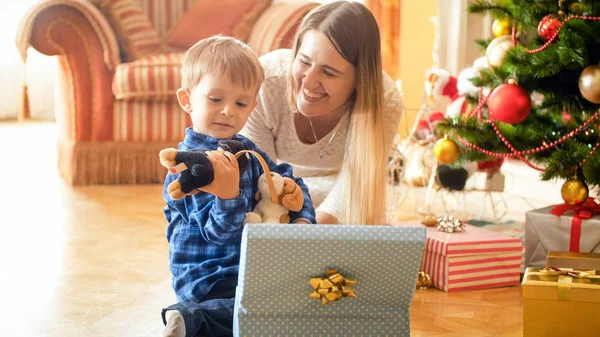 Портрет смеющейся молодой матери с маленьким улыбающимся мальчиком, открывающим рождественские подарки — стоковое фото