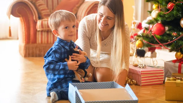 Очаровательный маленький мальчик сидит под елкой и обнимает плюшевую игрушку, которую он получил в подарочной коробке от Санты — стоковое фото