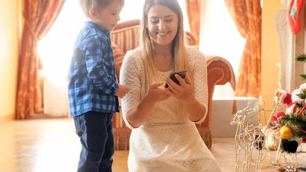 Πορτρέτο του χαμογελώντας νεαρή μητέρα δείχνει κινητό τηλέφωνο με τον μικρό γιο για πρωί των Χριστουγέννων — Φωτογραφία Αρχείου