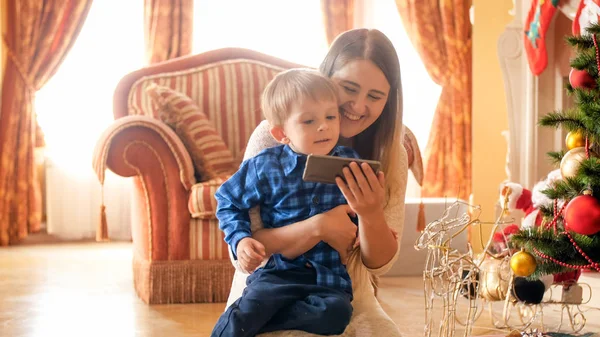 스마트 폰에 크리스마스 트리 및 보고 비디오에서 어머니와 함께 앉아 밝은 유아 소년 미소의 초상화 — 스톡 사진