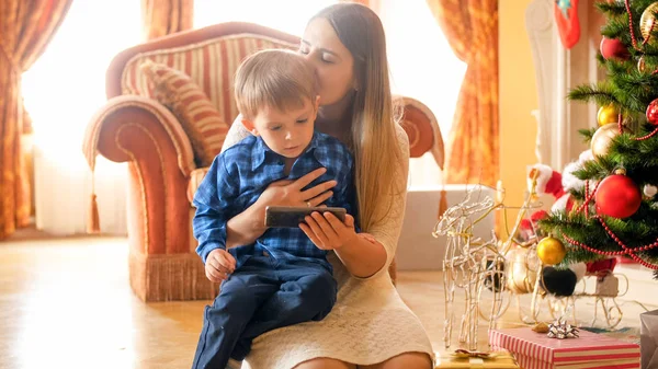 Hermosa madre joven besando a su hijo pequeño mientras él está viendo el video en el teléfono inteligente en la mañana de Navidad —  Fotos de Stock