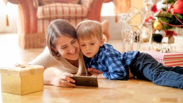 Retrato de mãe feliz sorridente com o menino deitado na sala de estar sob a árvore de Natal e assistindo vídeo no smartphone — Fotografia de Stock