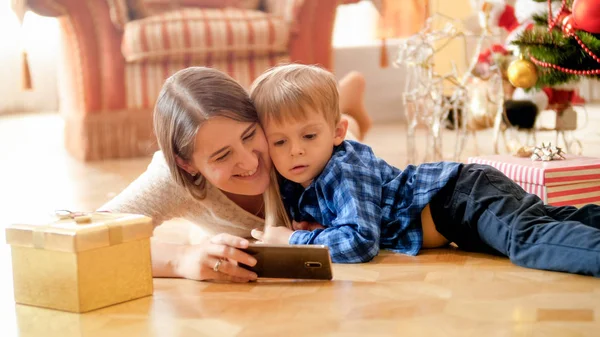 Porträtt av lilla pojken titta på tecknade serier på mobiltelefon med mamma under julgranen — Stockfoto