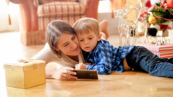 Retrato de bela jovem mãe deitada no chão com seu filho menor e assistindo desenhos animados no smartphone — Fotografia de Stock