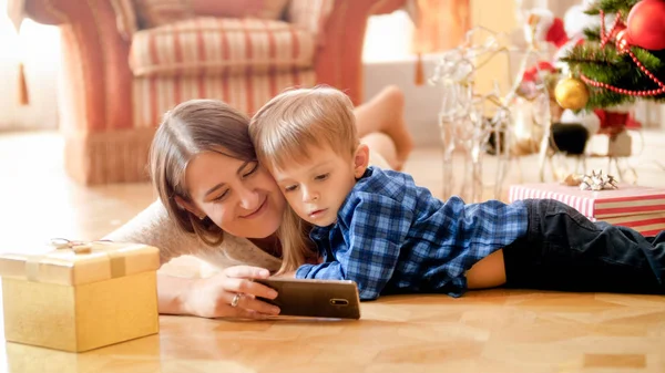 Jovem mãe gostando de assistir vídeo com o menino hertoddler no chão na sala de estar decorada para o Natal — Fotografia de Stock