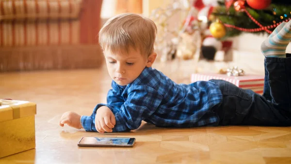 Ładny 3 lat toddler chłopiec leżącego pod choinkę i oglądać bajki na telefon komórkowy — Zdjęcie stockowe