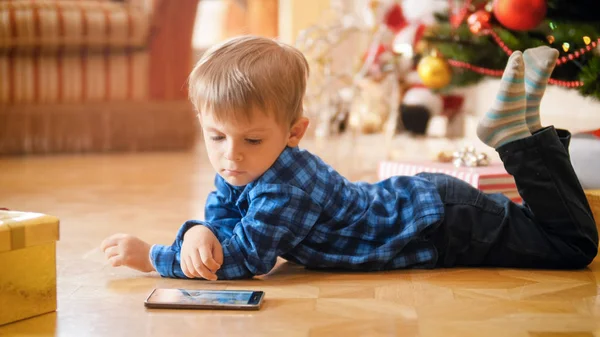 Kleiner Junge liegt unter Weihnachtsbaum und schaut Video auf Smartphone — Stockfoto