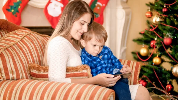 Bela jovem mãe sentada em poltrona com seu filhinho ao lado da árvore de Natal e assistindo vídeo no smartphone — Fotografia de Stock