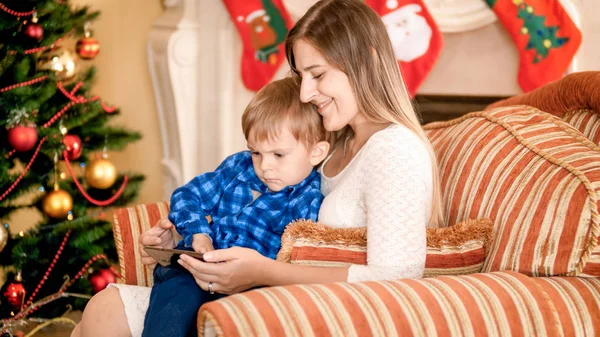 快乐微笑的母亲与她的孩子看视频在智能手机在扶手椅旁边的圣诞树 — 图库照片