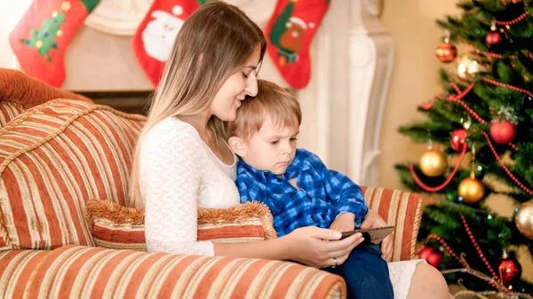 Anneyle Noel ağacının yanındaki oturma ve izlerken küçük yürümeye başlayan çocuk çizgi film üstünde hareket eden telefon — Stok fotoğraf