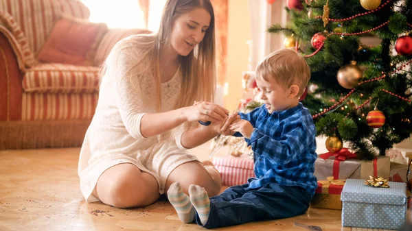 Hermosa madre joven sentada bajo el árbol de Navidad con su pequeño hijo y dándole confeti colorido —  Fotos de Stock