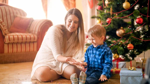 Glad skrattande småbarn pojke med vacker ung mamma på golvet täckt med färgglada konfetti bredvid granen — Stockfoto