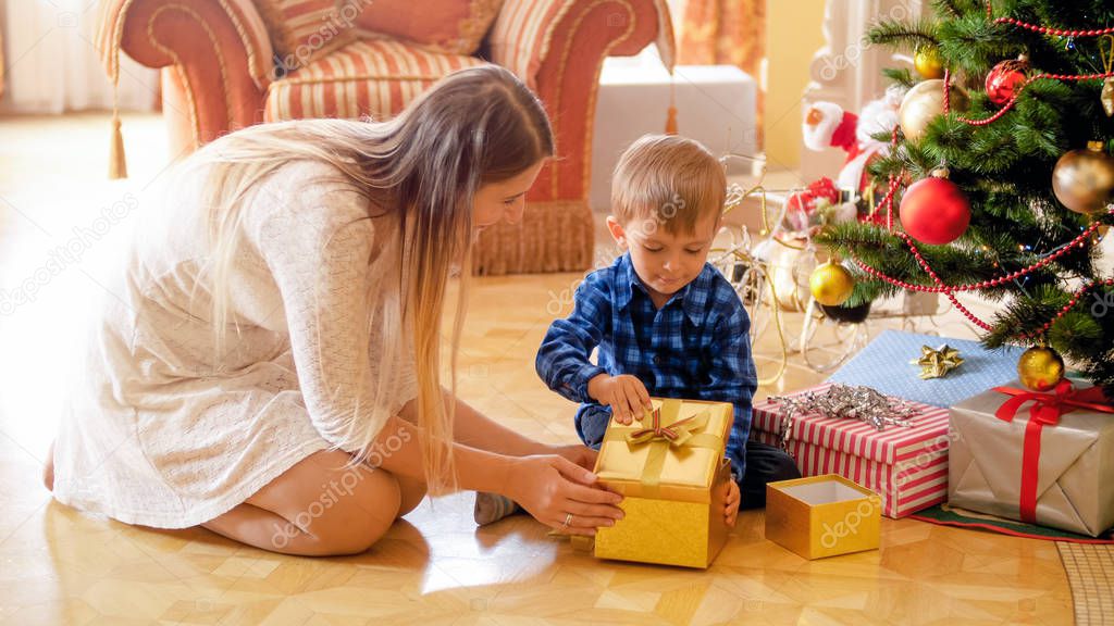 Cute little boy with mother sitting under Christmas tree and open gift box