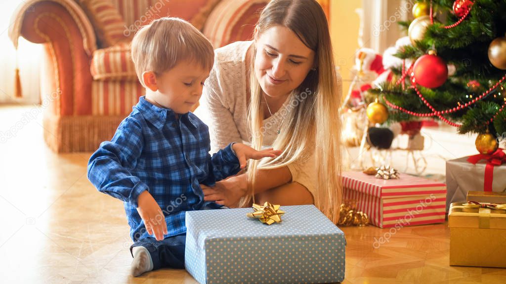 Portrait of young mother sitting with toddler boy under Christmas tree and looking at gift box with golden ribbon bow