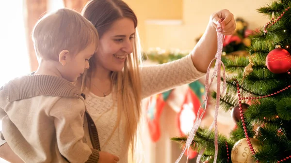 Porträtt av glada unga mor med liten pojke dekorera julgran med färgglada band — Stockfoto