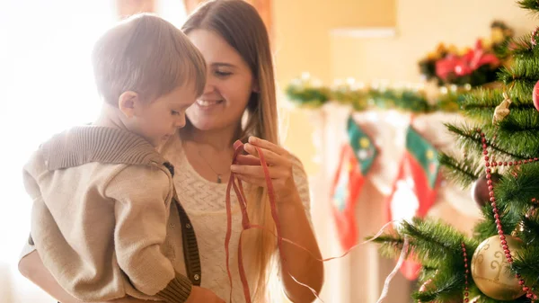 Bela sorridente jovem mãe abraçando o menino ao lado da árvore de Natal na sala de estar — Fotografia de Stock