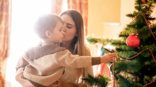 Mulher bonita beijando seu filho enquanto ele está decorando a árvore de Natal — Fotografia de Stock