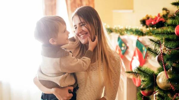 Retrato de una joven madre sonriente mirando a su pequeño hijo en la sala de estar decorada para Navidad —  Fotos de Stock