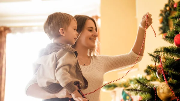 Anya díszítő karácsonyfa színes gyöngyök, mosolygós kisfiú portréja — Stock Fotó