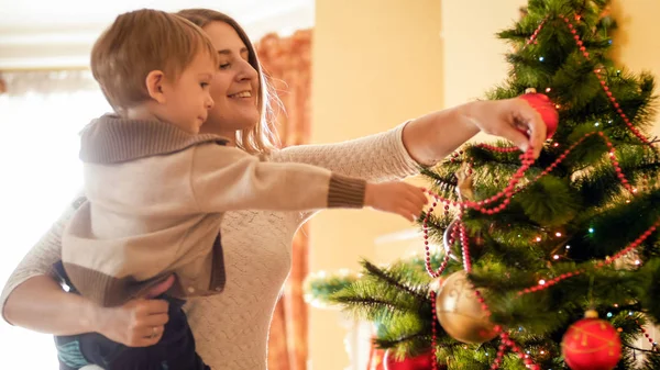 Retrato de primer plano de la alegre madre oyung con el niño pequeño decorando el árbol de Navidad con cuentas brillantes rojas —  Fotos de Stock