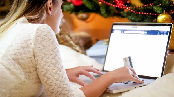 Closeup image of young woman with credit card lying under Christmas tree and ordering gifts for winter holidays — Stock Photo, Image