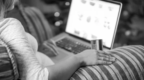 Primer plano imagen en blanco y negro de una joven sentada en un sillón con tarjeta de crédito y la tienda en línea de navegación en el ordenador portátil —  Fotos de Stock