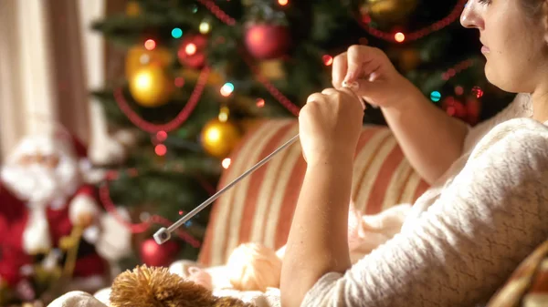 Imagen de cerca de una joven sentada junto al árbol de Navidad y tejiendo bufanda de lana — Foto de Stock