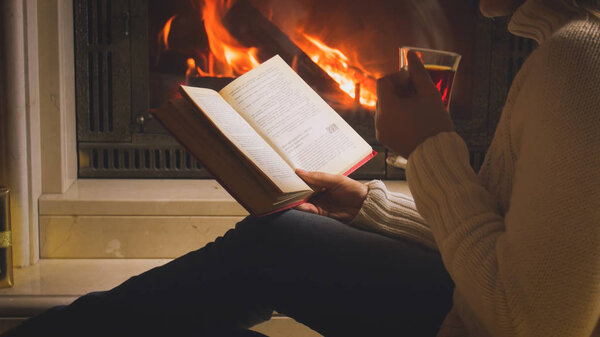 Closeup toned photo of woman with book drinking tea by the fireplace at night