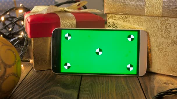 Closeup 4k footage of smartphone leaning on Christmas gift box and glowing light garland. Green chromakey display for inserting your image or video on screen — Stock Video