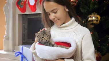 Noel ağacının yanındaki Noel Baba şapkalı oturan güzel genç kız sevişme şirin yavru kedi portresi