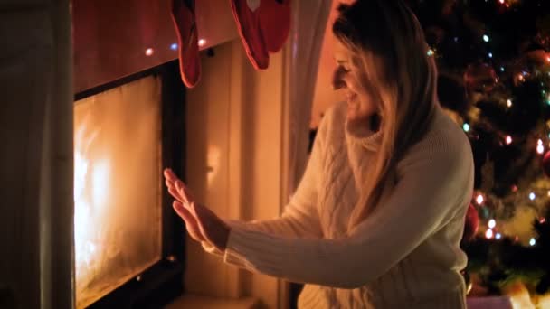 4k видео красивой улыбающейся женщины в шерстяном свитере, греющейся у горящего камина ночью — стоковое видео