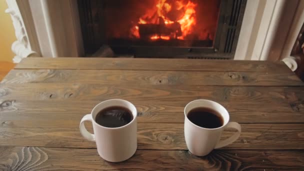 Zbliżenie zwolnionym tempie nagrania dwóch kubków z gorącą herbatę na drewnianym stole obok spalania kominek w domu — Wideo stockowe
