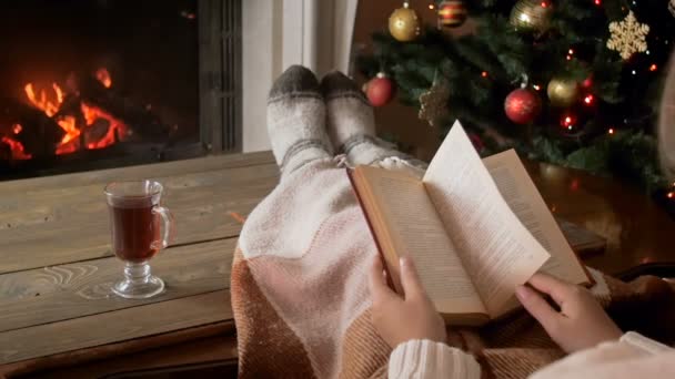 Медленное видео, где молодая женщина читает книгу на диване рядом с горящим камином и пьет чай в рождественскую ночь — стоковое видео