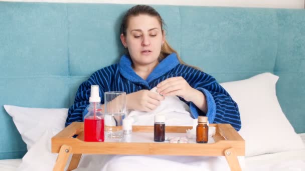 Imágenes de 4k de una mujer joven sintiéndose enferma acostada en la cama y usando muchos medicamentos. Mujer aplicando garganta y aerosol nasal — Vídeo de stock