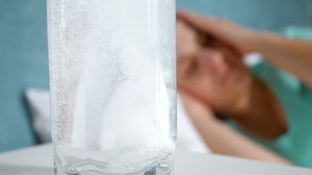 Closeup vídeo em câmera lenta de pílula de aspirina caindo em vidro de água e dissolvendo. Mulher deitada na cama e sofrendo de dor de cabeça — Vídeo de Stock