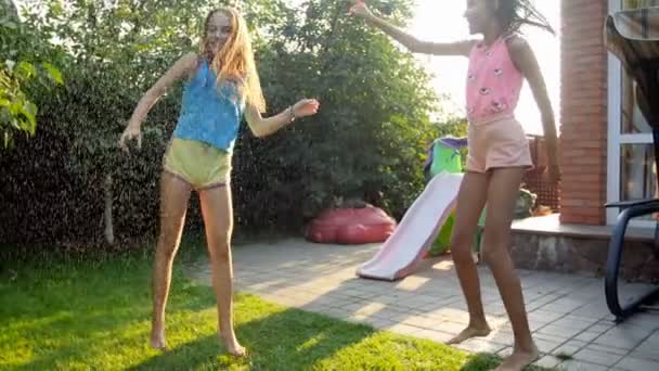 Slow motion beelden van twee tienermeisjes dansen en springen onder warme zomerregen op achtertuin lachen — Stockvideo