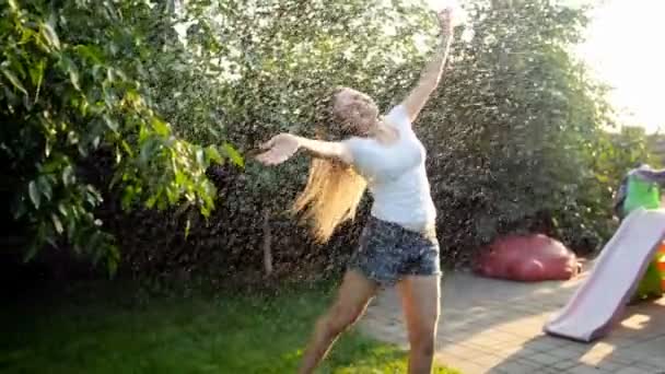 Slow-motion video van gelukkig vrolijke jonge vrouw met lange haren dansen onder zomerregen in achtertuin — Stockvideo