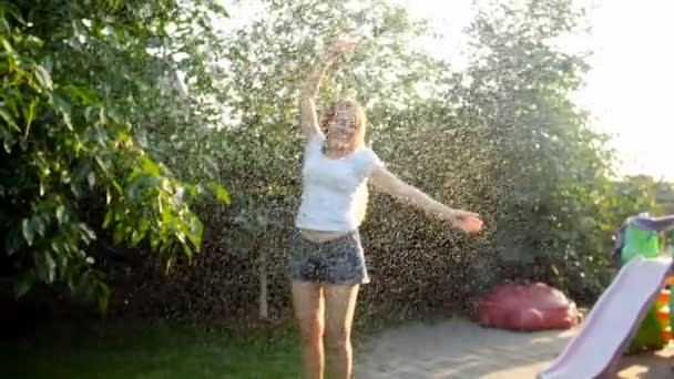 Imágenes en cámara lenta de una hermosa joven sonriente disfrutando de la lluvia de verano al atardecer en el patio trasero — Vídeos de Stock