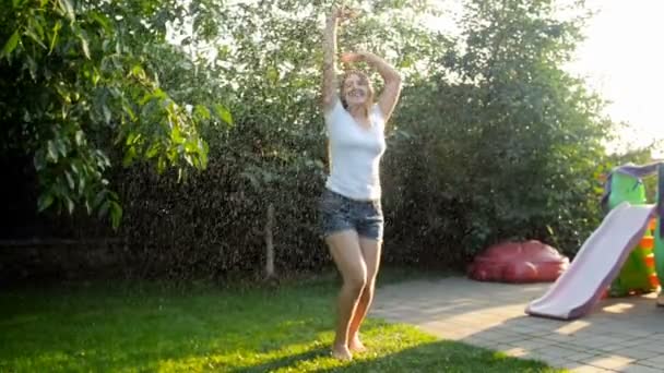 Slow-motion video van lachen jonge vrouw dansen en springen onder water van tuin sprinkler bij zonsondergang — Stockvideo