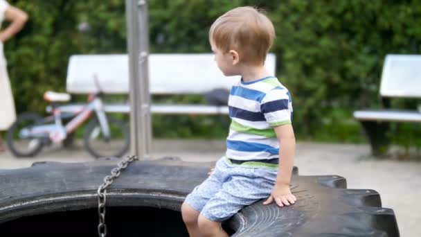 4k 视频可爱的微笑学步的小男孩坐在大轮胎在公园操场上 — 图库视频影像