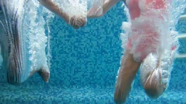 Unterwasseraufnahmen in Zeitlupe zeigen zwei Mädchen, die händchenhaltend in ein Schwimmbad springen und unter Wasser tauchen. Luftblasen heben sich langsam an. — Stockvideo